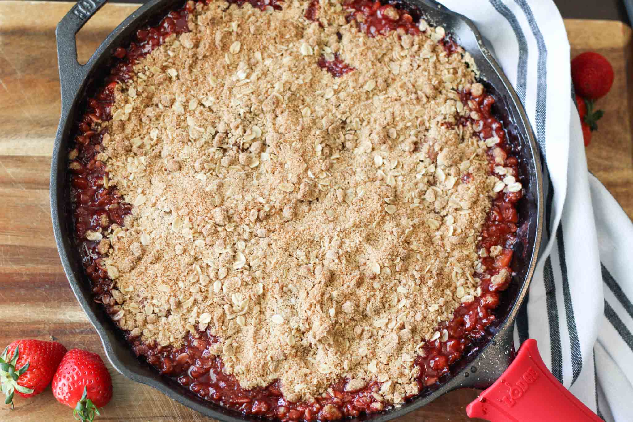 finished strawberry-rhubarb crisp cooked in cast iron skillet on wood cutting board with whole strawberries as decoration.