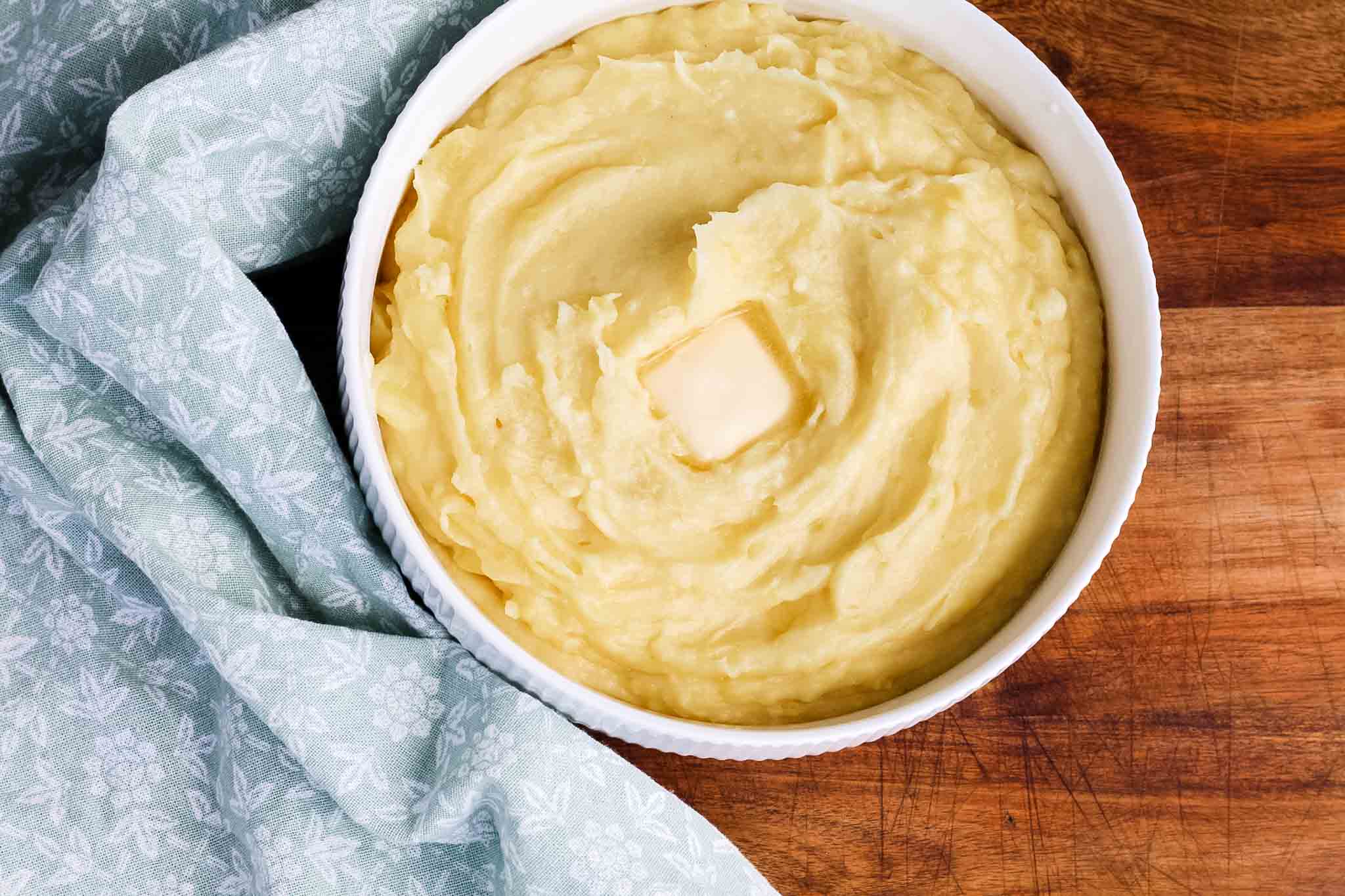 white bowl of mashed potatoes on wood cutting board with pad of butter on top